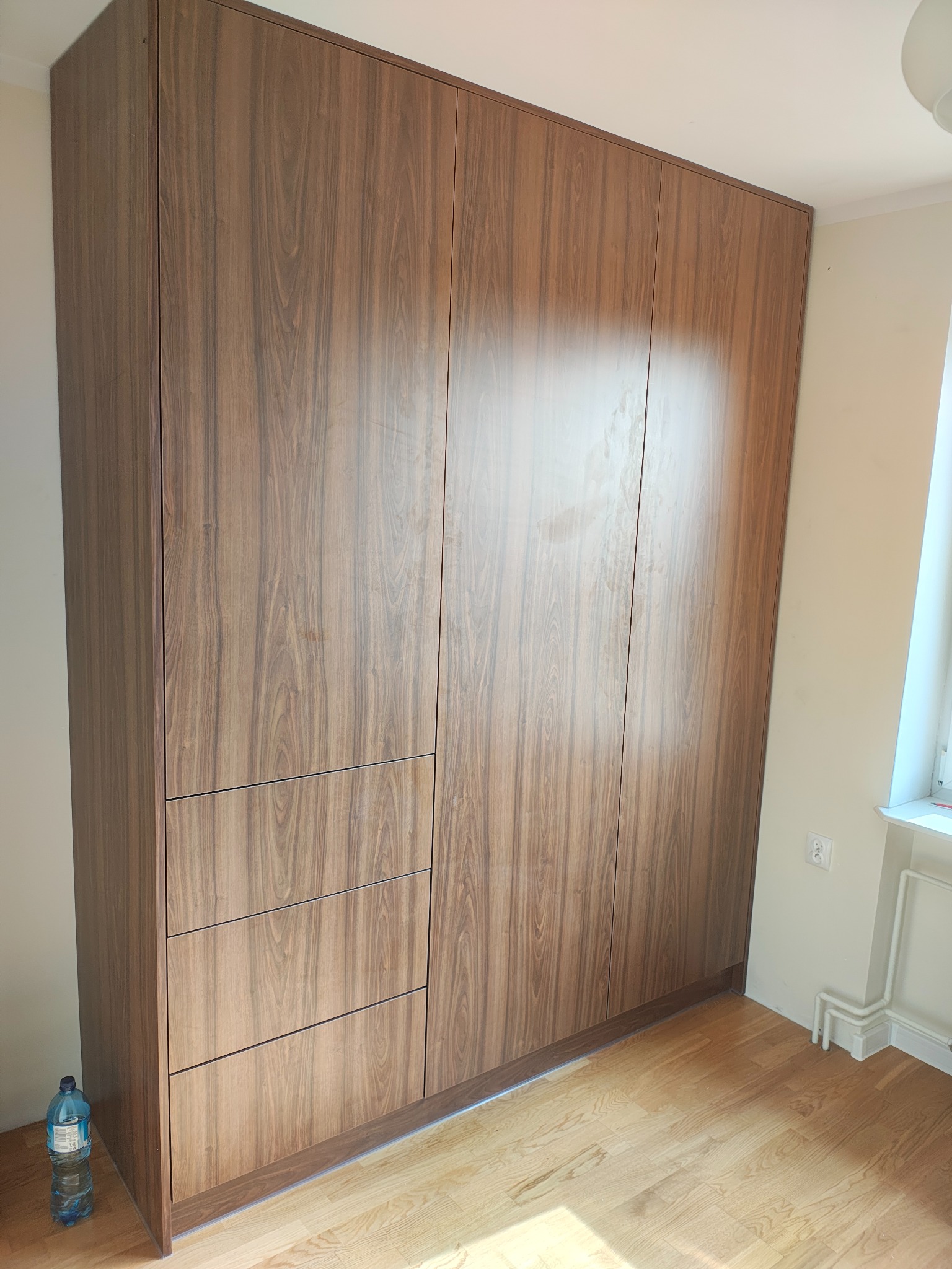 Wysoka, zabudowana szafa wnękowa z drewnopodobnymi frontami, podzielona na segment z trzema szufladami i część drzwiową. Butelka wody przy podstawie szafy.