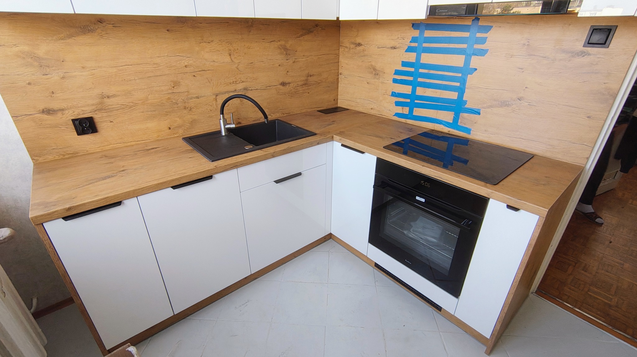 Kompaktowa kuchnia w kształcie litery L z białymi frontami i blatem z drewna o naturalnym odcieniu. Widoczny zlew, płyta indukcyjna i piekarnik.