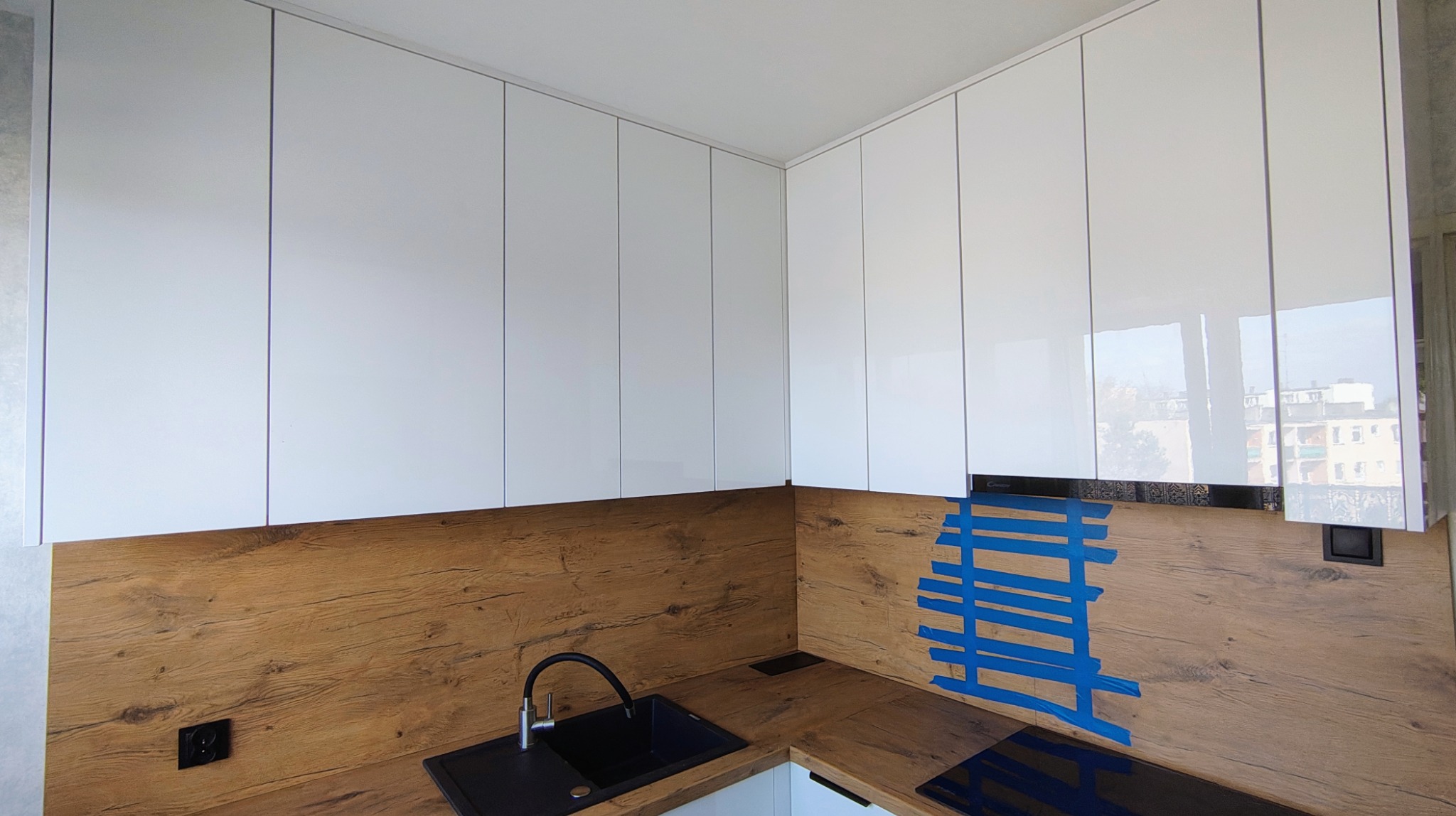 Białe, lakierowane szafki kuchenne podwieszane nad blatem z okleiną drewnopodobną. Widoczny fragment zlewu i płyty indukcyjnej. Niebieska taśma maskująca na ścianie.
