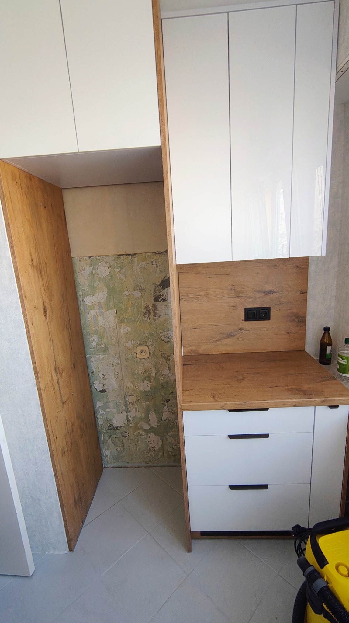 Montaż białej kuchni z drewnianymi akcentami. Widoczna ściana w trakcie remontu, kontrastująca z nowymi meblami i czarnymi uchwytami szuflad.