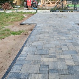 Tom-Bruk - Ułożona, szara kostka brukowa z czarnym obrzeżem na posesji. W tle widać ogrodzenie i elementy placu budowy. Osoba na paletach po lewej stronie kadru.