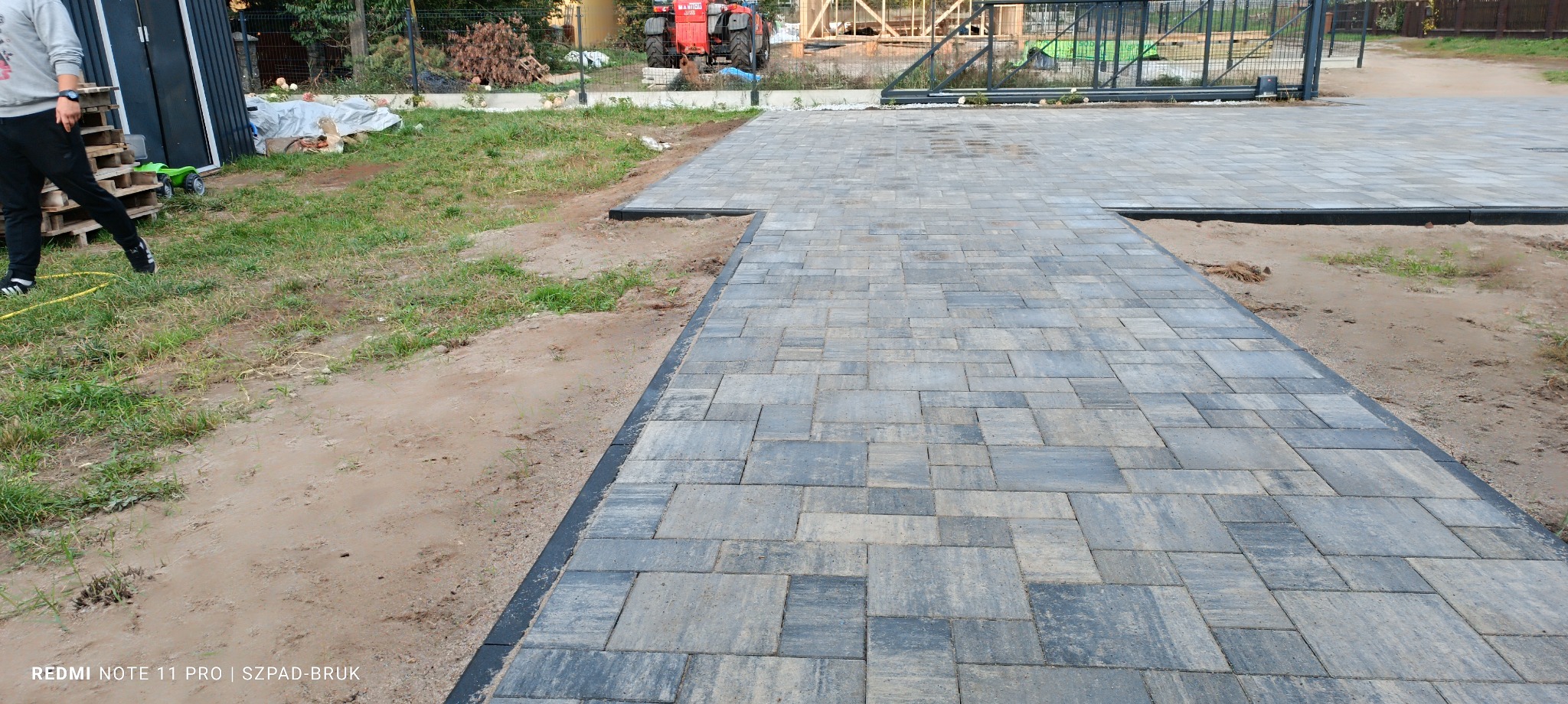 Ułożona, szara kostka brukowa z czarnym obrzeżem na posesji. W tle widać ogrodzenie i elementy placu budowy. Osoba na paletach po lewej stronie kadru.