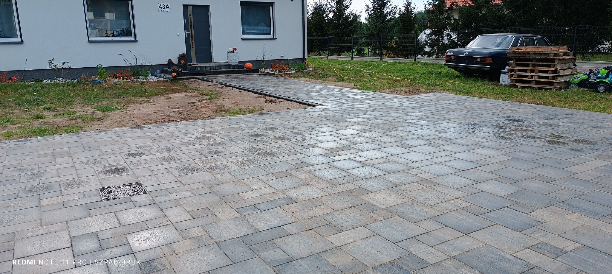 Ułożona, szara kostka brukowa na podjeździe domu jednorodzinnego z numerem 43a. Widoczny właz kanalizacyjny i obrzeża. W tle auto i palety.
