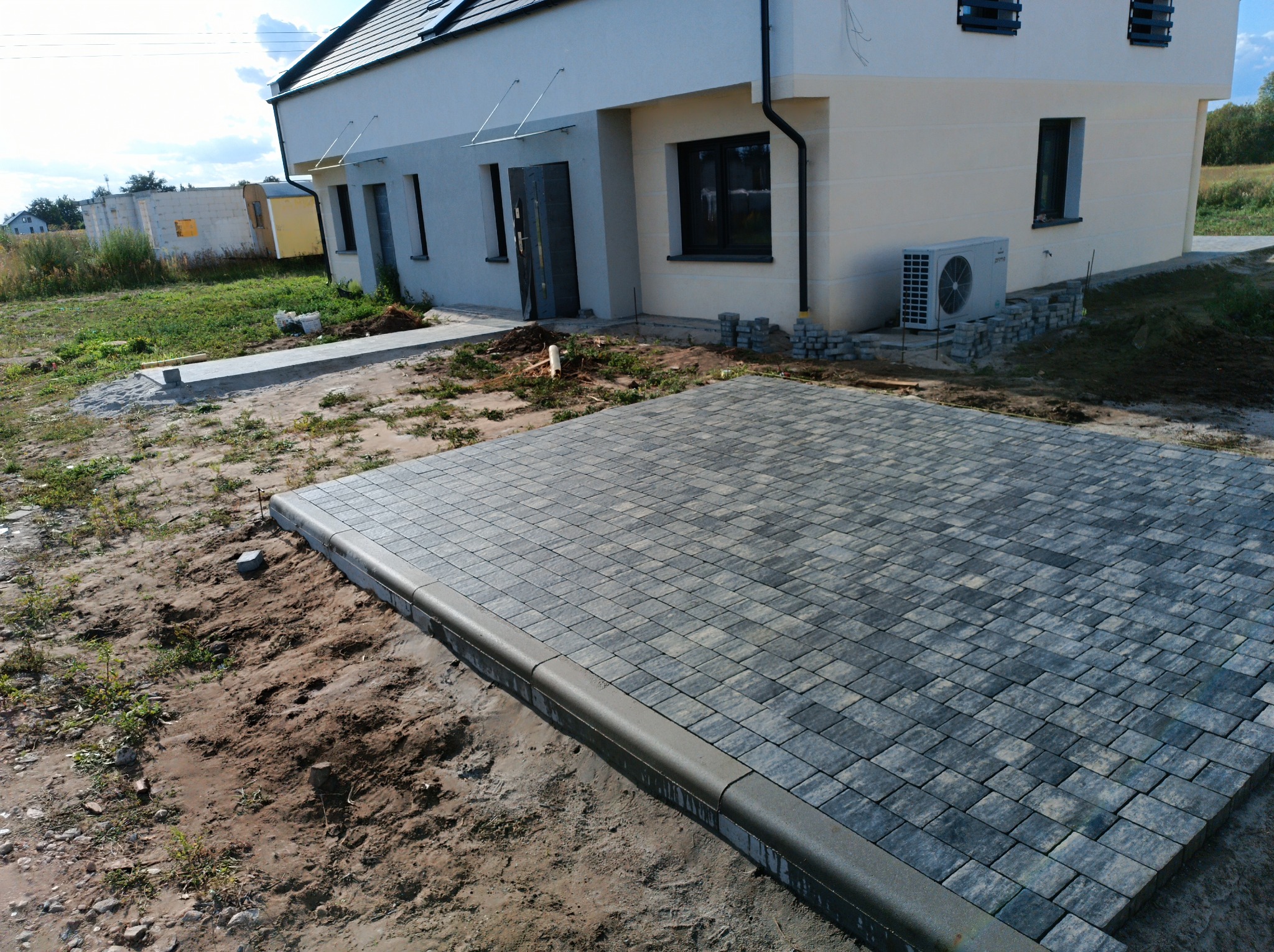 Ułożona kostka brukowa w szaro-niebieskich odcieniach przed nowym domem. Widoczne obrzeża i przygotowany teren pod dalsze prace brukarskie.