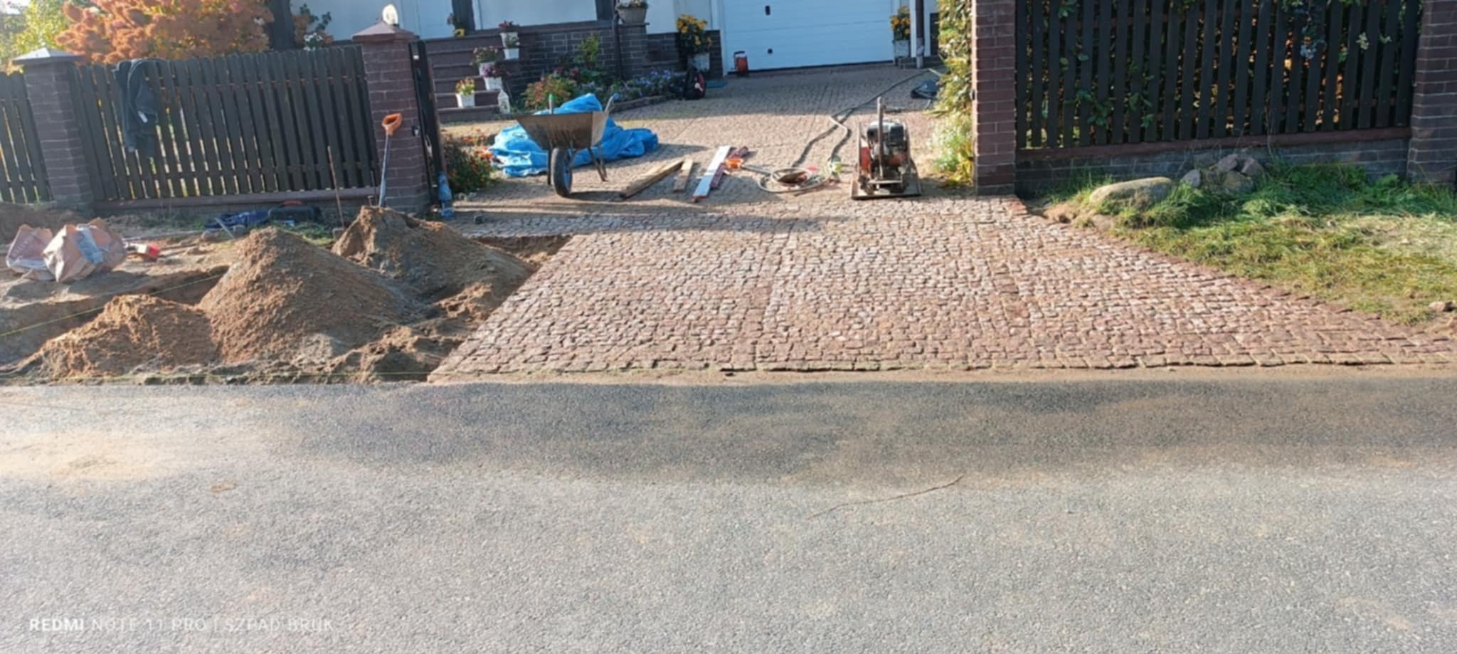Częściowo ułożona kostka granitowa na podjeździe, z widoczną stertą piasku i narzędziami, sugerująca trwające prace brukarskie.