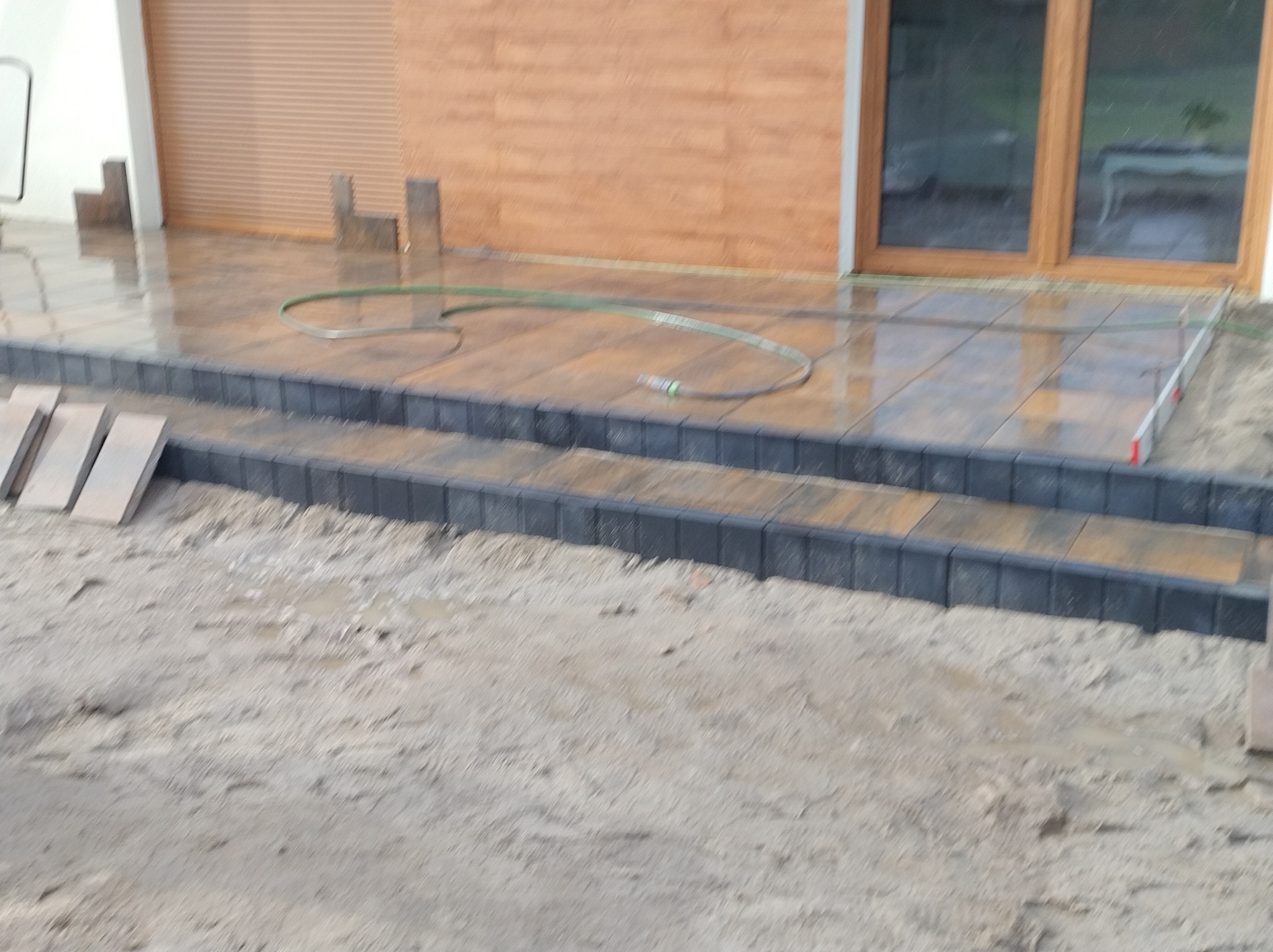 Ułożona, mokra kostka brukowa na tarasie z dwoma stopniami, widoczna woda i wąż ogrodowy. Częściowo widoczny teren przed tarasem.