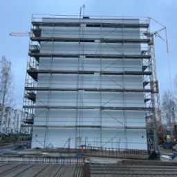 Budynek w trakcie ocieplania elewacji, widoczne rusztowania i częściowo ocieplona ściana. Na pierwszym planie zbrojenie. W tle dźwig i drzewa.