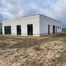 Zakład Budowlany ''Kocięda'' Stefan Kocięda - Nowoczesny, parterowy dom z szarą elewacją i czarnymi oknami; widoczny fragment placu budowy i pracująca osoba w oddali. Pochmurne niebo w tle.