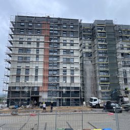 Zakład Budowlany ''Kocięda'' Stefan Kocięda - Wielopiętrowy budynek w trakcie prac elewacyjnych, otoczony rusztowaniami. Widoczni pracownicy w kaskach, sprzęt budowlany i częściowo ukończona elewacja w odcieniach szarości i brązu.