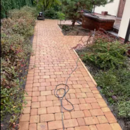 Ułożona, brązowo-pomarańczowa kostka brukowa tworząca ścieżkę w ogrodzie, widoczna zagęszczarka w tle i kabel elektryczny na kostce.