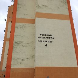 Zakład Budowlany ''Kocięda'' Stefan Kocięda - Wielopiętrowy budynek z pomarańczowo-kremową elewacją, widoczne anteny na dachu. Ślady zabrudzeń na jasnej części fasady. Tablica z napisem 'Wspólnota Mieszkaniowa'.