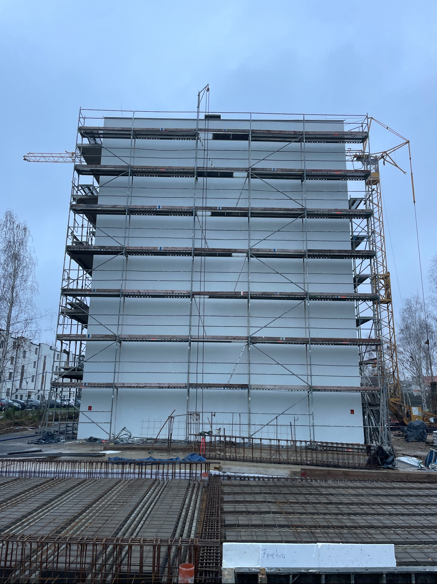 Budynek w trakcie ocieplania elewacji, widoczne rusztowania i częściowo ocieplona ściana. Na pierwszym planie zbrojenie. W tle dźwig i drzewa.