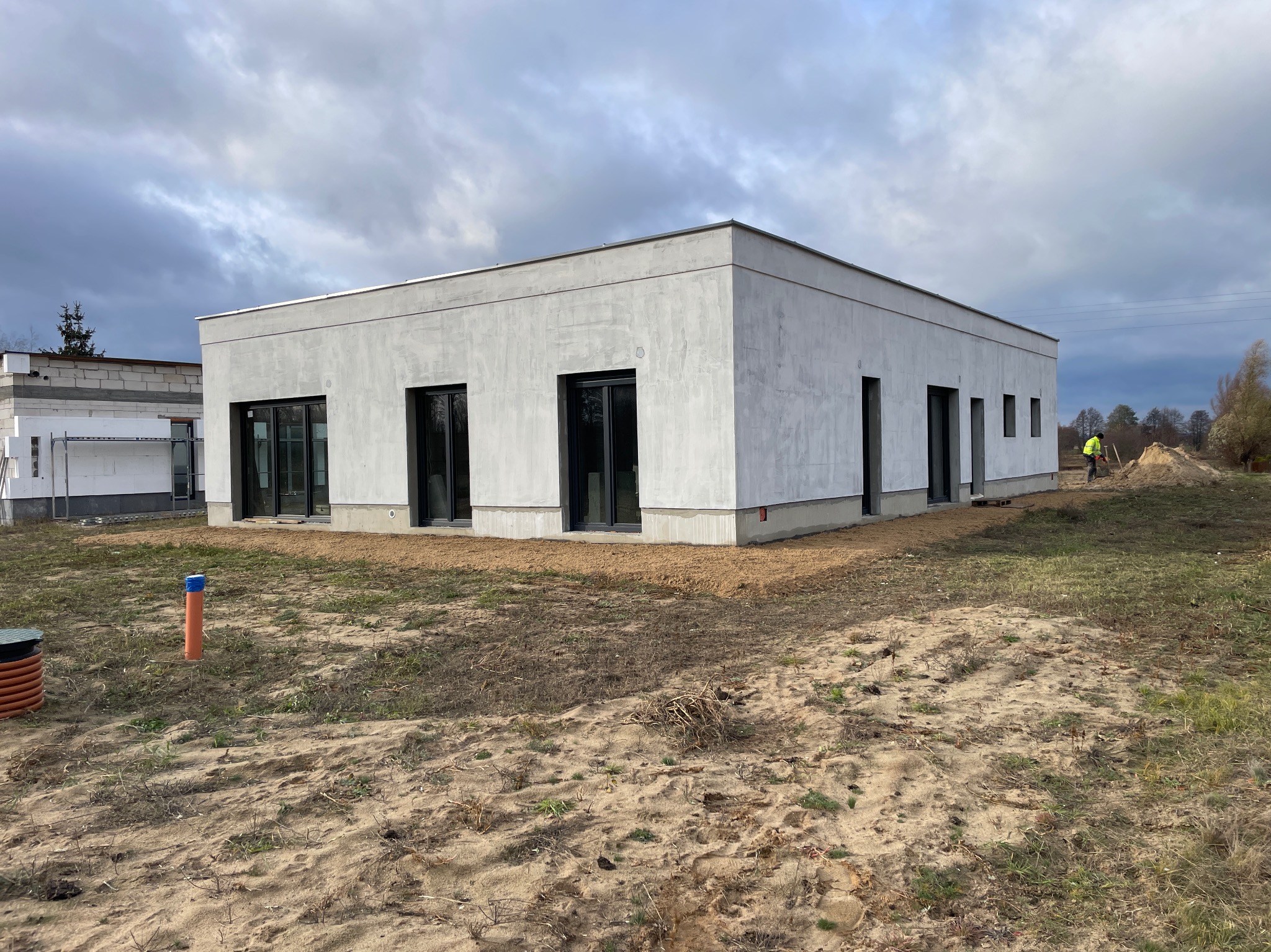 Nowoczesny, parterowy dom z szarą elewacją w trakcie wykończenia. Widoczne czarne okna i drzwi, piaskowe podłoże i pracownik w oddali.