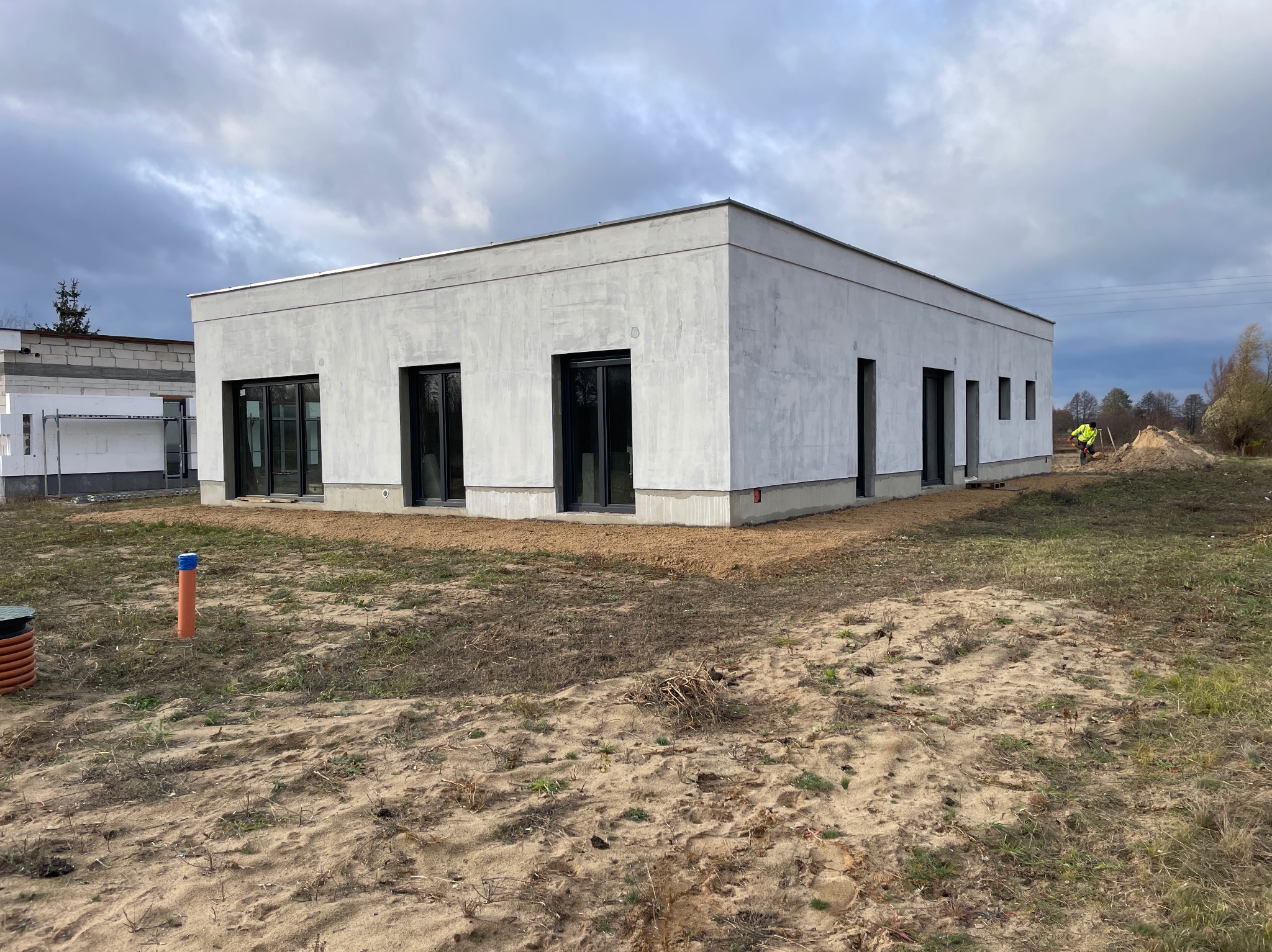 Nowoczesny, parterowy dom z szarą elewacją i czarnymi oknami; widoczny fragment placu budowy i pracująca osoba w oddali. Pochmurne niebo w tle.