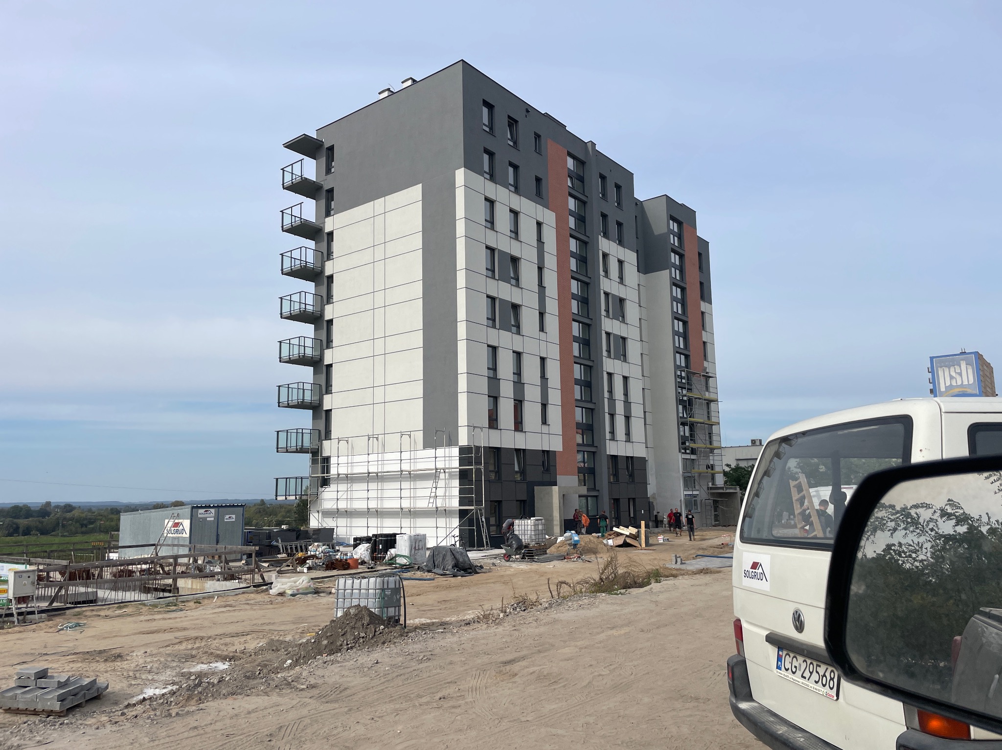 Wielopiętrowy budynek w trakcie prac elewacyjnych, widoczne rusztowania i ekipa budowlana. Częściowo ocieplona fasada, biała furgonetka z boku kadru.