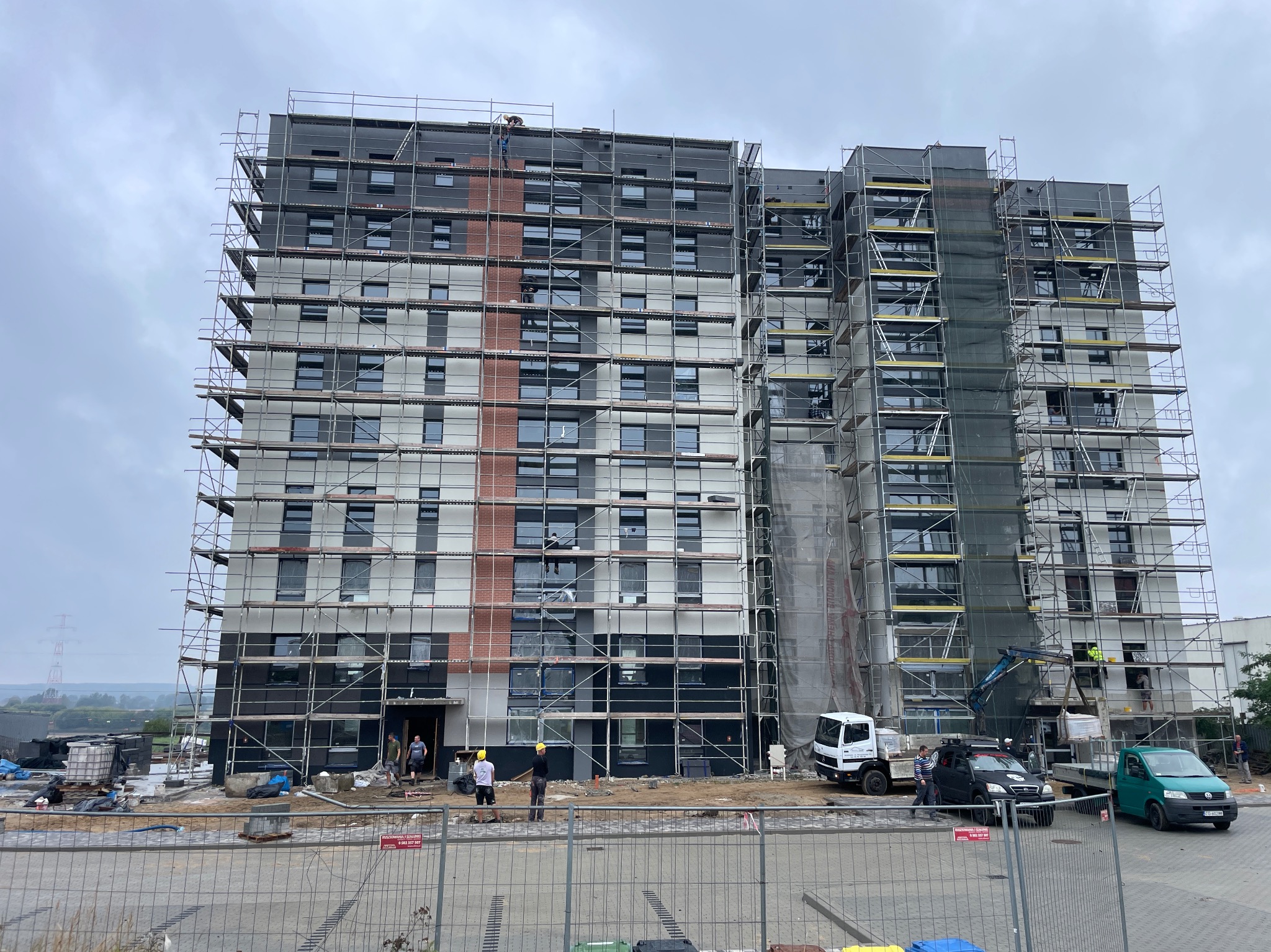 Wielopiętrowy budynek w trakcie prac elewacyjnych, otoczony rusztowaniami. Widoczni pracownicy w kaskach, sprzęt budowlany i częściowo ukończona elewacja w odcieniach szarości i brązu.