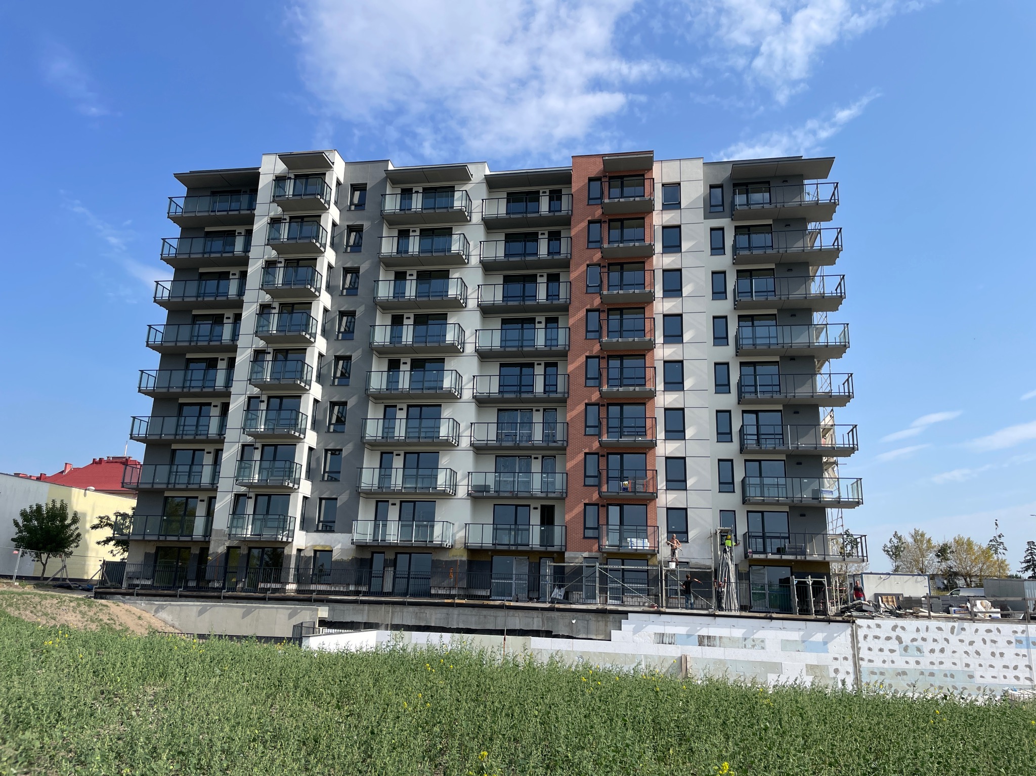 Wielopiętrowy budynek z balkonami w trakcie prac elewacyjnych. Widoczne rusztowania i pracownicy. Część elewacji szara, część biała z elementami ceglanymi.