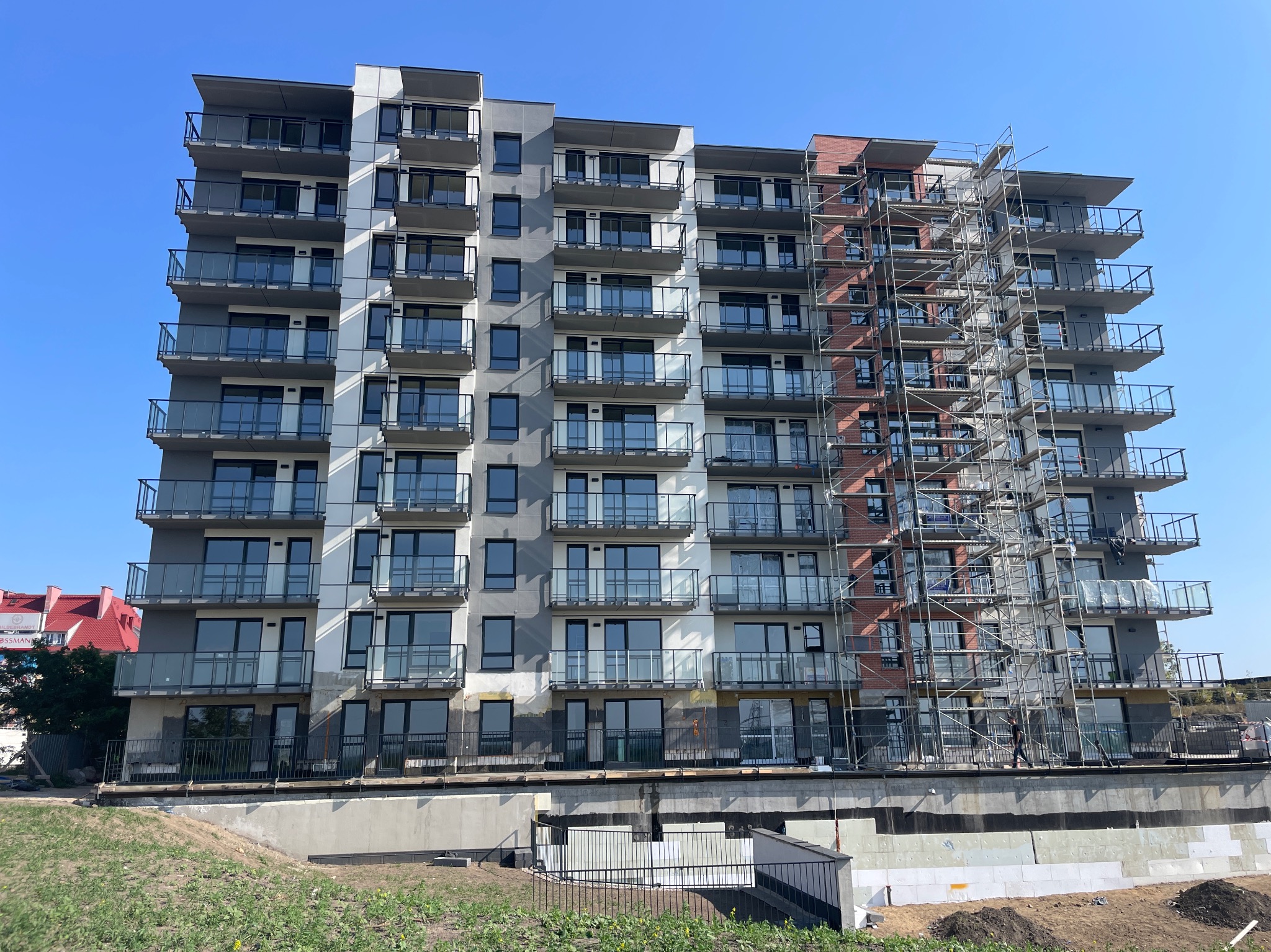 Wielopiętrowy budynek z rusztowaniami na bocznej ścianie. Widoczne balkony z szklanymi balustradami. Część elewacji w szarych odcieniach, fragment w cegle.