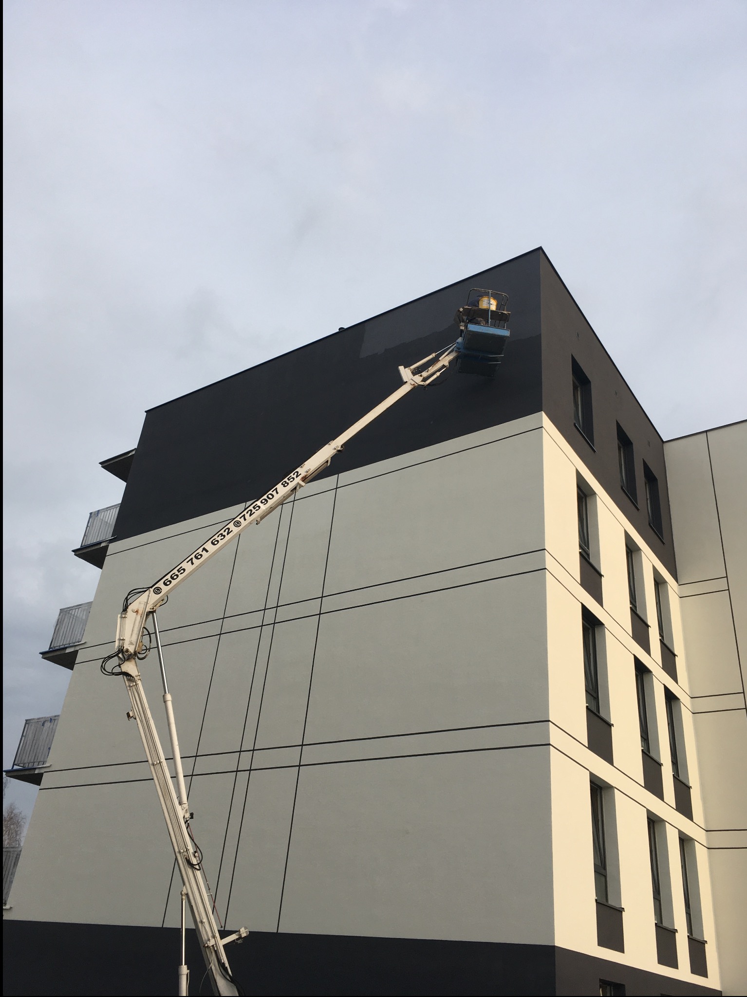 Malowanie elewacji bloku mieszkalnego z użyciem podnośnika koszowego. Nowoczesna architektura, dwukolorowa fasada. Widoczne numery kontaktowe na ramieniu podnośnika.