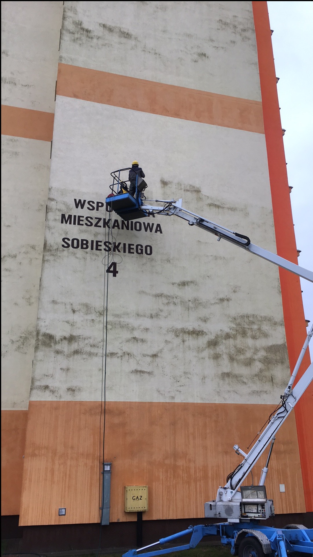 Pracownik na podnośniku czyści elewację budynku z napisem 'Wspólnota Mieszkaniowa Sobieskiego 4'. Widoczne zabrudzenia na ścianie, żółta skrzynka gazowa.