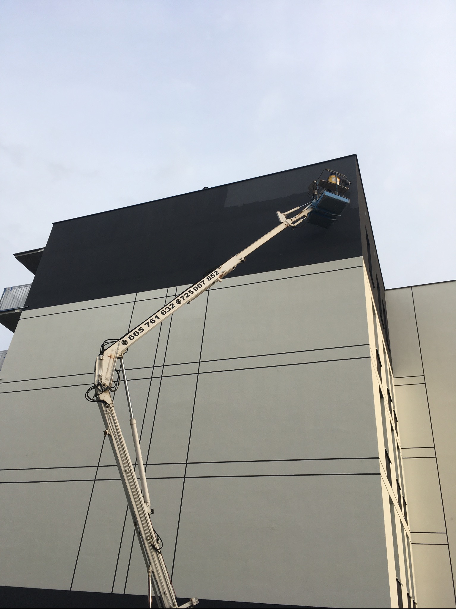 Prace na wysokościach. Podnośnik koszowy przy czarno-białej elewacji budynku. Widoczny numer telefonu na ramieniu podnośnika. Prace elewacyjne w toku.