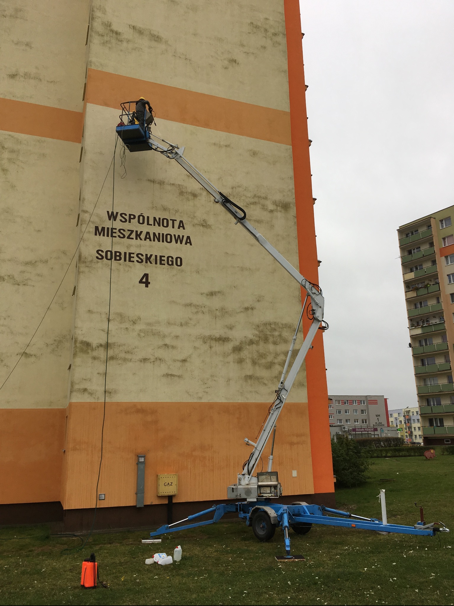 Pracownik na podnośniku czyści elewację bloku mieszkalnego z napisem 'Wspólnota Mieszkaniowa Sobieskiego 4', widoczne ślady zabrudzeń i butelki z płynem czyszczącym.