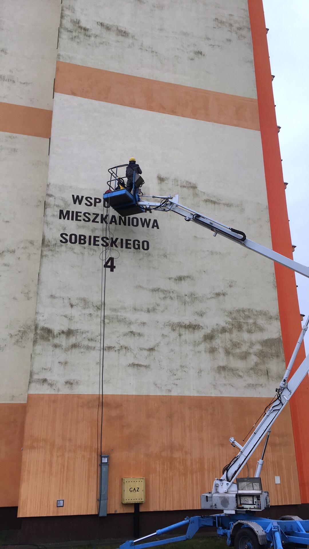 Pracownik na podnośniku czyści elewację budynku z napisem 'Wspólnota Mieszkaniowa Sobieskiego 4'. Widoczne zabrudzenia na ścianie i pomarańczowe pasy.