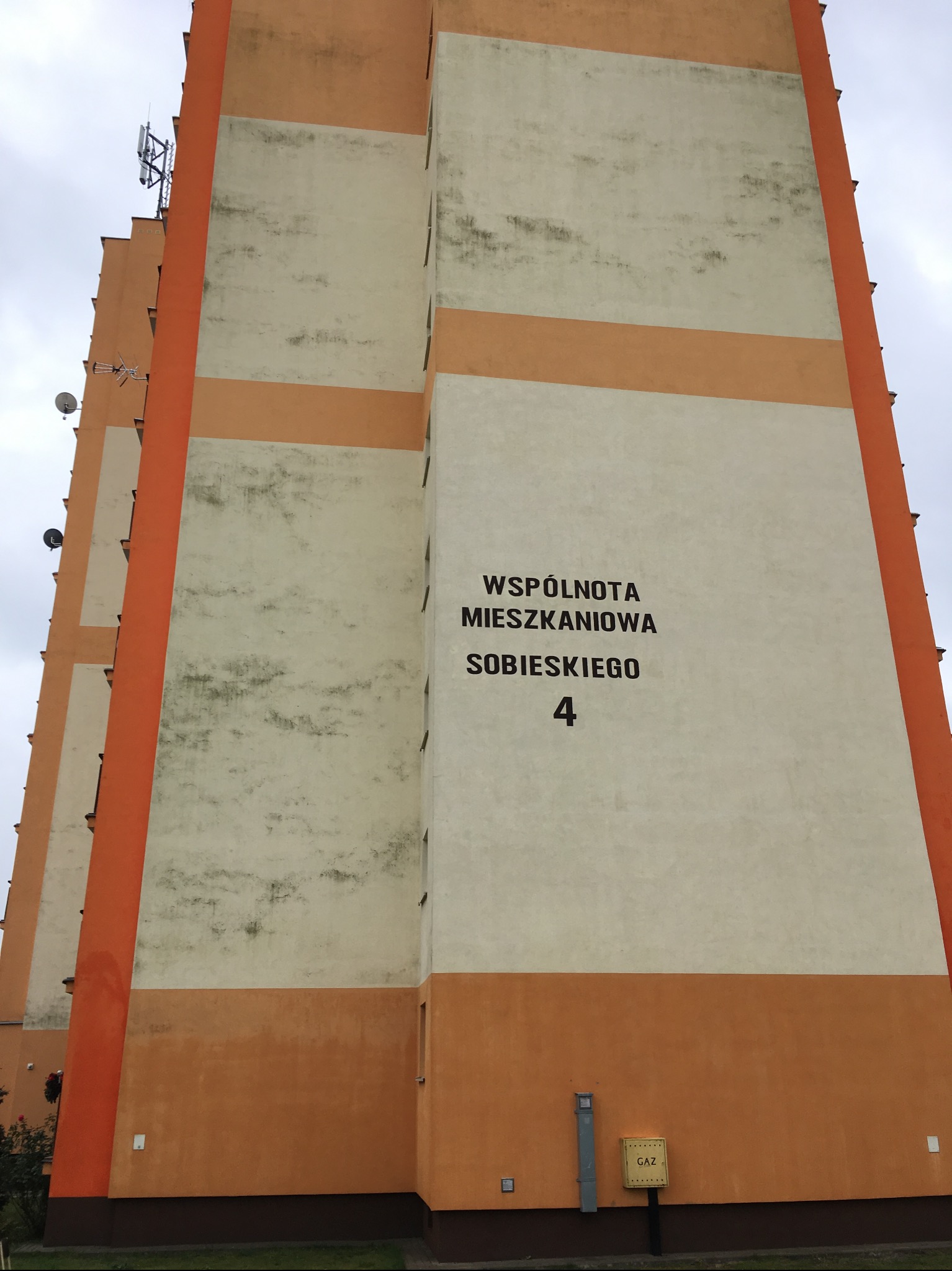 Wielopiętrowy budynek z pomarańczowo-kremową elewacją, widoczne anteny na dachu. Ślady zabrudzeń na jasnej części fasady. Tablica z napisem 'Wspólnota Mieszkaniowa'.