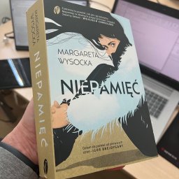 MW Studio Projekt - Marta Wróblewska - Książka Margarety Wysockiej 'Niepamięć' trzymana w dłoni na tle biurka z laptopem. Okładka z ilustracją kobiety i tytułem. Widoczny bok książki z nazwiskiem autorki.