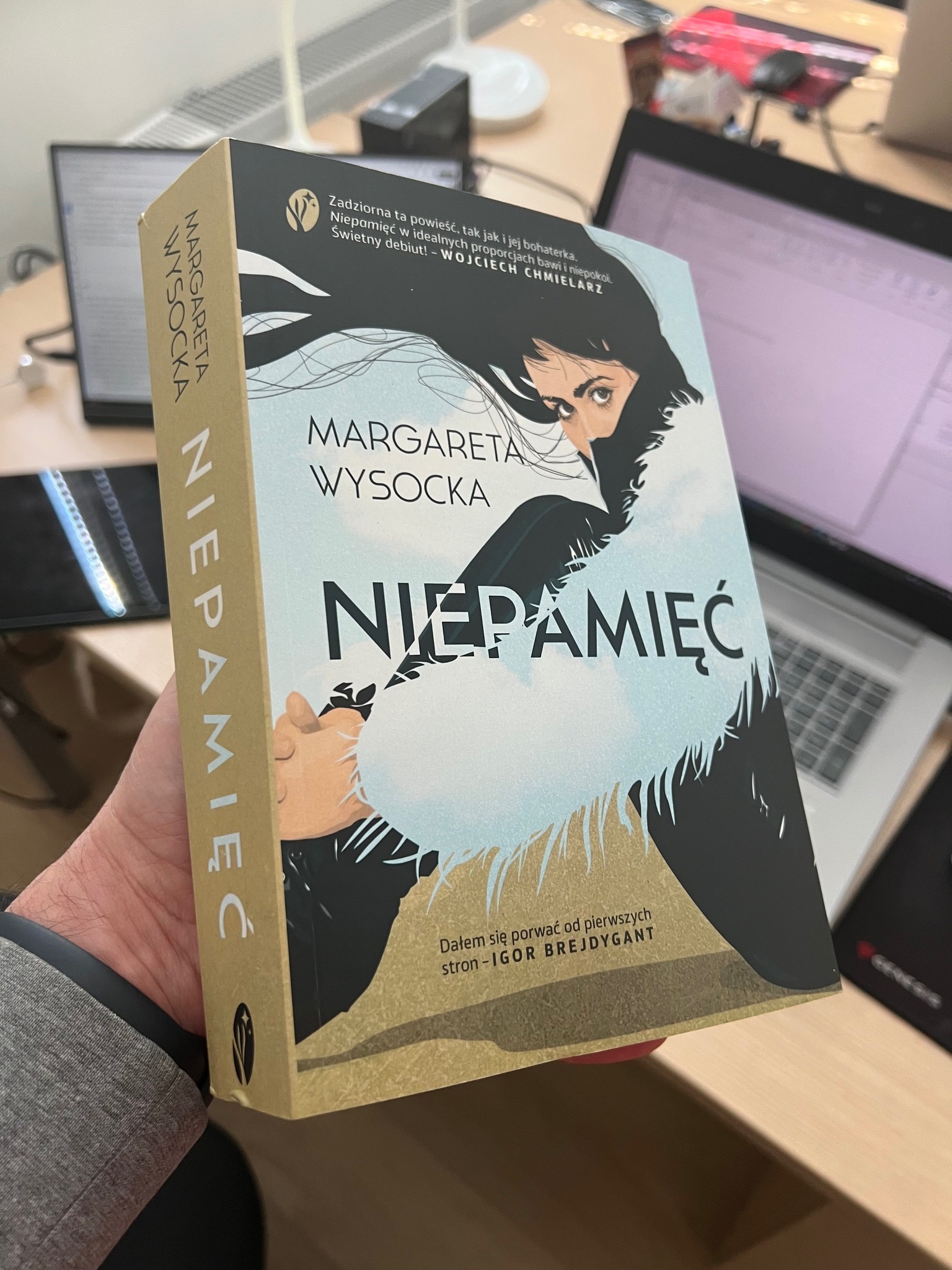 Książka Margarety Wysockiej 'Niepamięć' trzymana w dłoni na tle biurka z laptopem. Okładka z ilustracją kobiety i tytułem. Widoczny bok książki z nazwiskiem autorki.