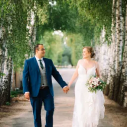 Para młoda spaceruje aleją brzóz, trzymając się za ręce. Panna młoda w białej sukni z bukietem, pan młody w niebieskim garniturze. Uśmiechnięci, w naturalnym oświetleniu.