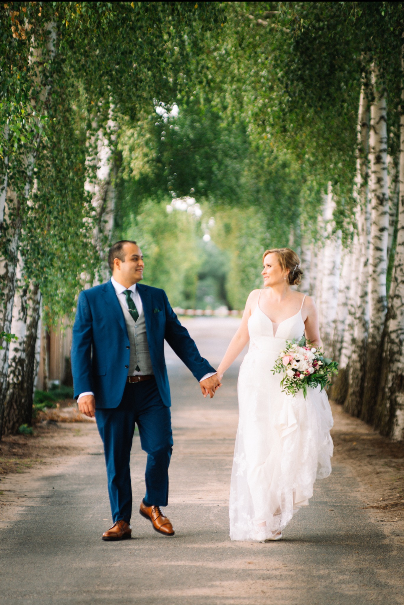 Para młoda spaceruje aleją brzóz, trzymając się za ręce. Panna młoda w białej sukni z bukietem, pan młody w niebieskim garniturze. Uśmiechnięci, w naturalnym oświetleniu.