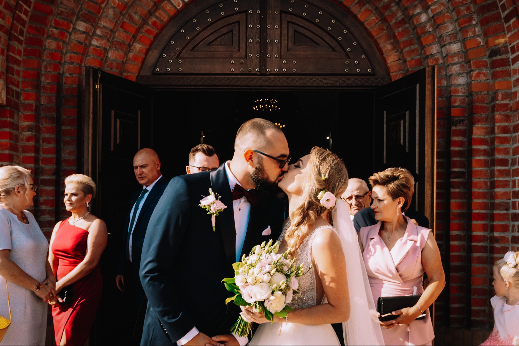 Para młoda w pocałunku przed ceglanym budynkiem, goście weselni w tle. Panna młoda trzyma bukiet. Ujęcie w słoneczny dzień.
