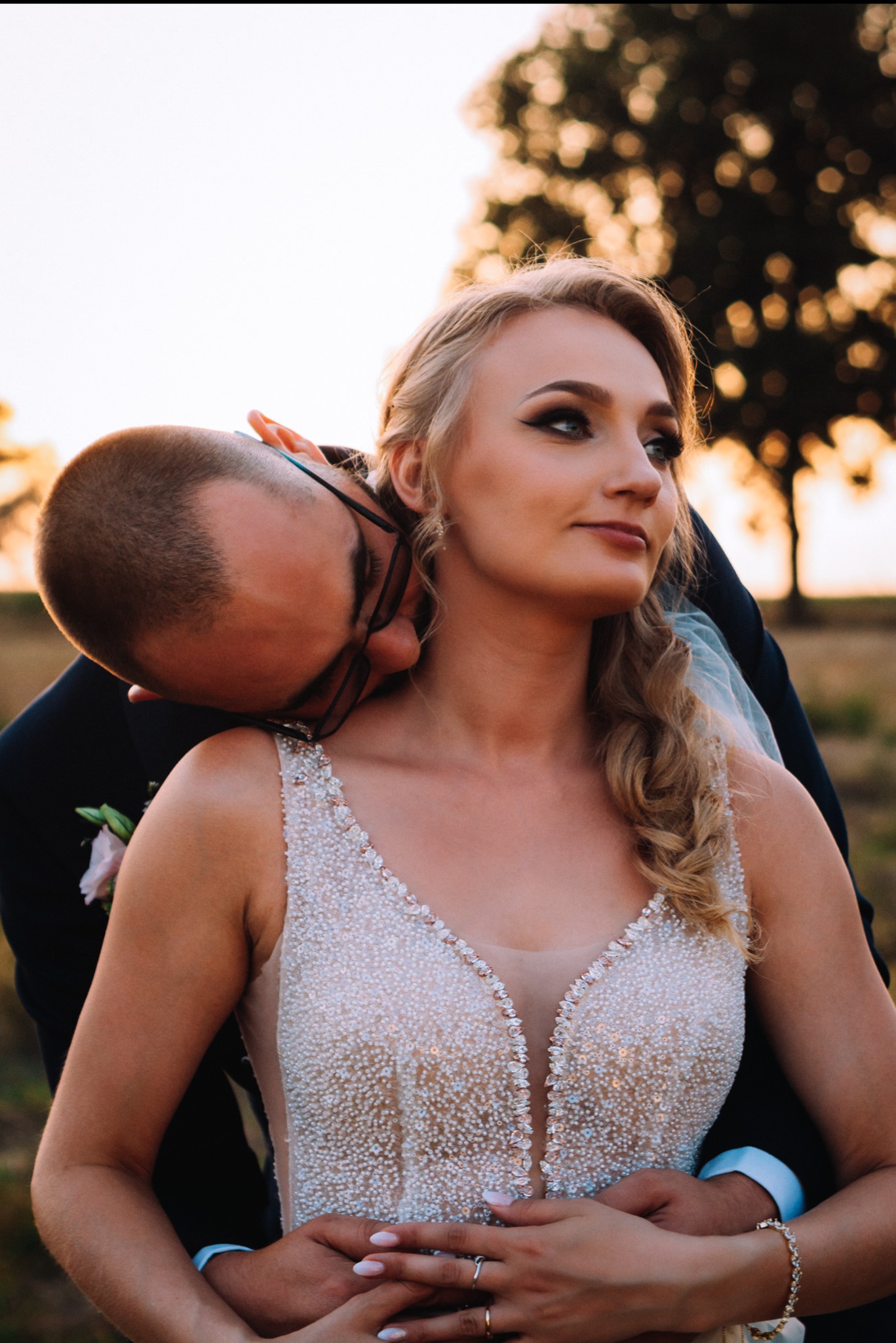 Ujęcie pary młodej w plenerze: panna młoda w zdobionej sukni spogląda w bok, obejmowana przez pana młodego w garniturze, z drzewem w tle.