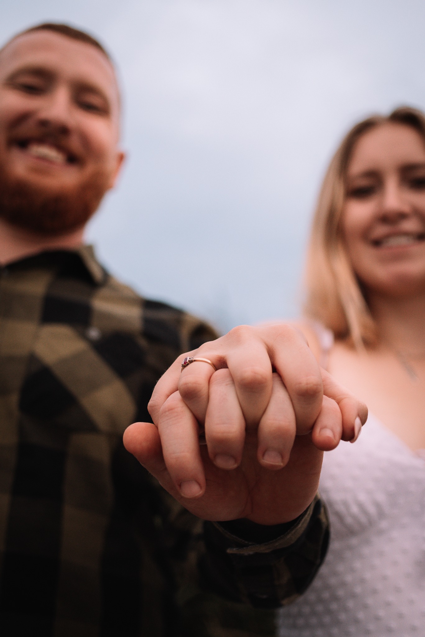 Zbliżenie splecionych dłoni pary z pierścionkiem zaręczynowym, uśmiechnięci, w tle niebo. Ujęcie z dołu podkreśla bliskość i intymność chwili.