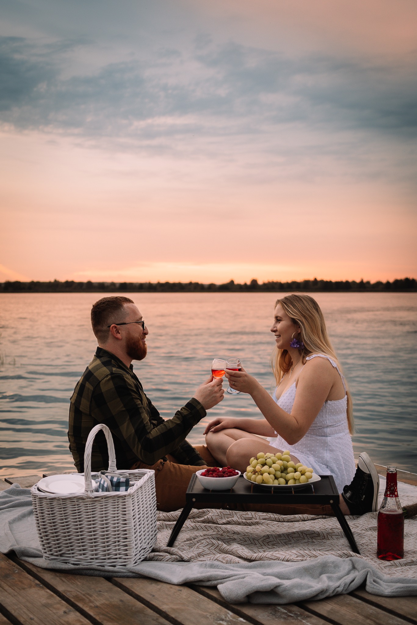 Romantyczny piknik nad wodą w Kazimierzu Biskupim: para wznosi toast kieliszkami wina o zachodzie słońca. Kosz piknikowy, owoce i butelka napoju na drewnianym pomoście.