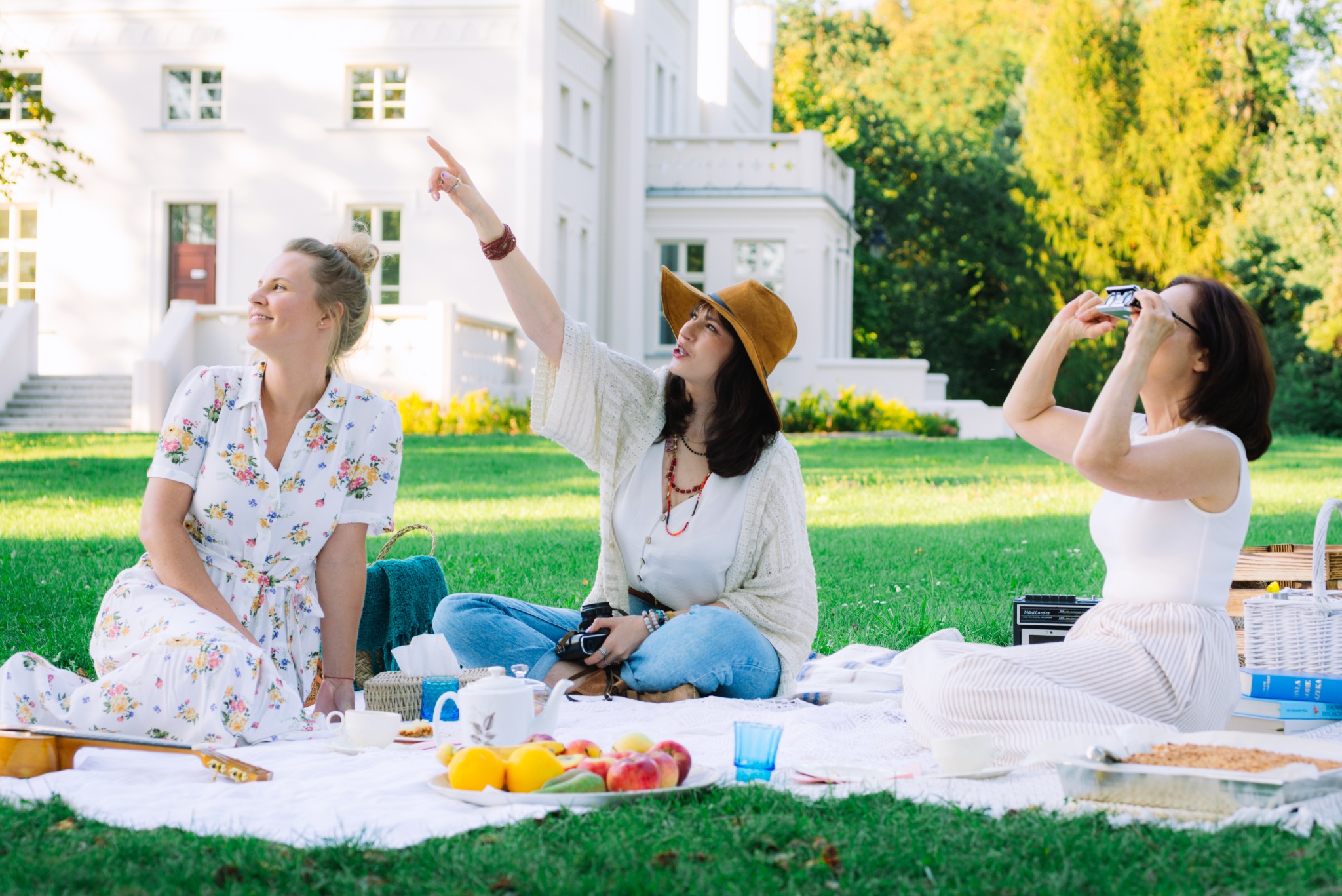 Trzy kobiety na pikniku na trawie przed białym budynkiem. Jedna fotografuje, druga wskazuje palcem, trzecia siedzi z uśmiechem. Owoce, gitara i kosz piknikowy.