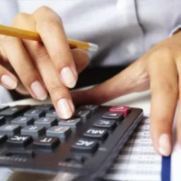 Kalkulator w użyciu: dłoń z oł&oacute;wkiem naciska przycisk na kalkulatorze, widoczne dokumenty z liczbami w tle. Blat biurka i fragment koszuli.