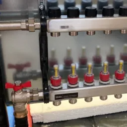 Rozdzielacz ogrzewania podłogowego TECE z zaworami, widok z bliska. Elementy instalacji zamontowane na styropianie, rury doprowadzające wodę, estetyczne wykonanie.