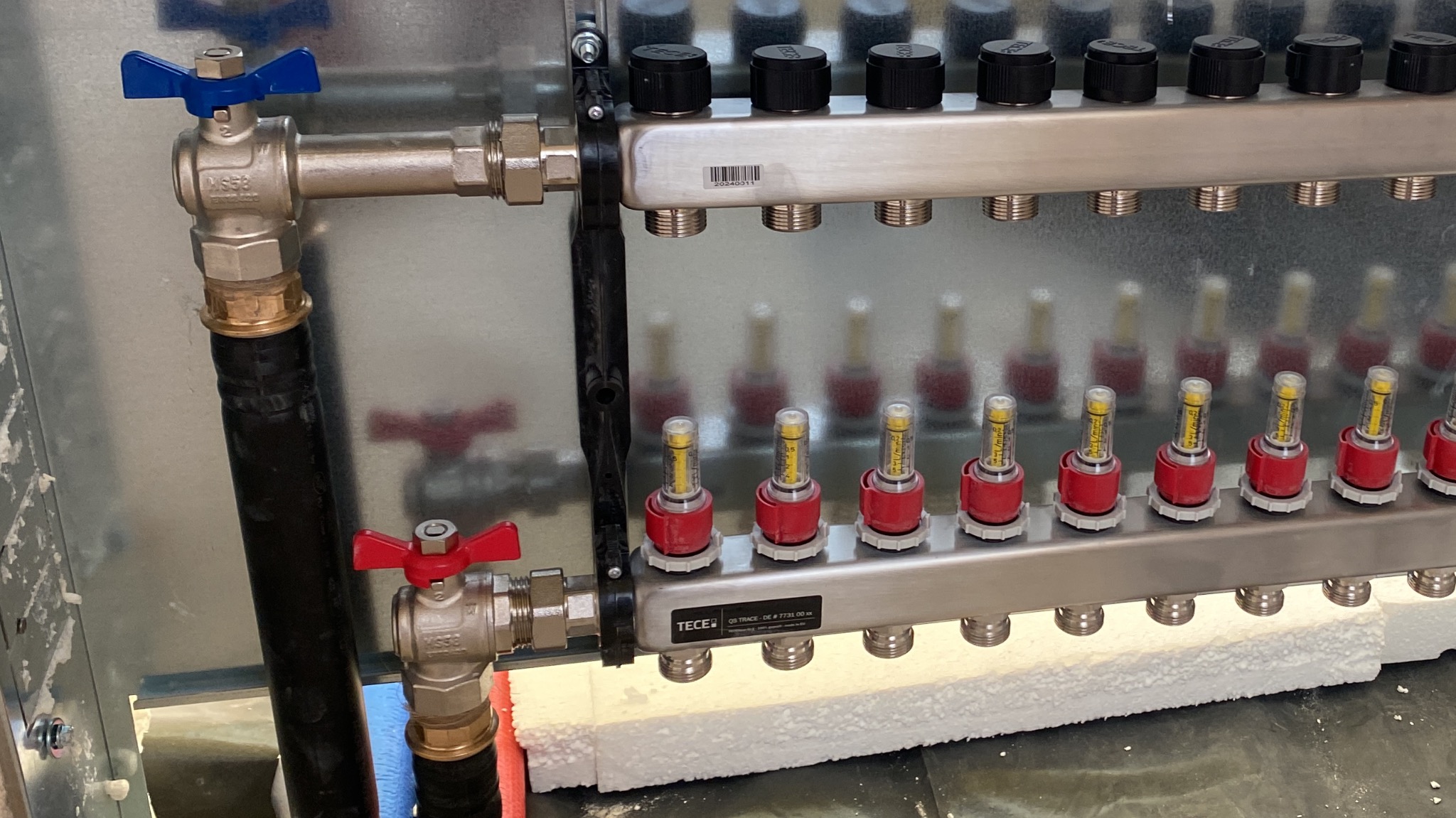 Rozdzielacz ogrzewania podłogowego TECE z zaworami, widok z bliska. Elementy instalacji zamontowane na styropianie, rury doprowadzające wodę, estetyczne wykonanie.