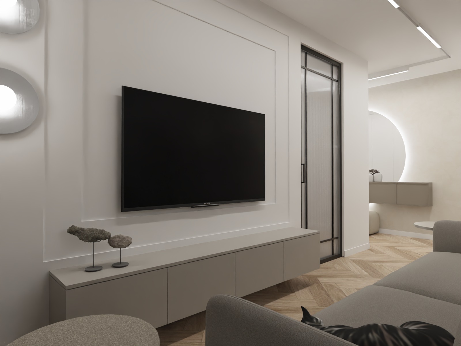 Minimalistyczny salon z telewizorem na ścianie, szarą szafką RTV i dekoracjami. Parkiet w jodełkę, okrągłe lustro z podświetleniem. W tle widoczny fragment sofy.