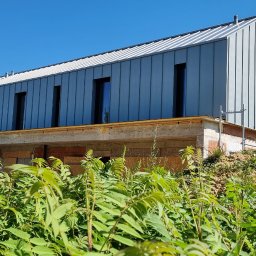 Blach-dek Mateusz Michalski - Nowoczesny dom z szarą blachą na dachu i elewacji, widoczny w letnim słońcu, z bujną zielenią na pierwszym planie. Budynek w trakcie budowy, widoczne rusztowania.