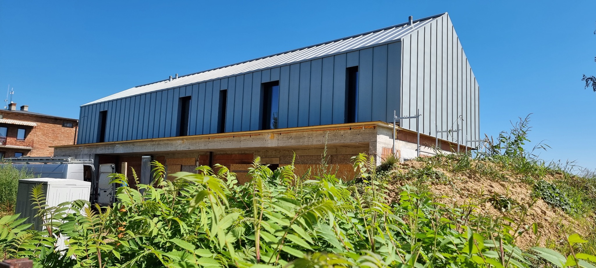 Nowoczesny dom z szarą blachą na dachu i elewacji, widoczny w letnim słońcu, z bujną zielenią na pierwszym planie. Budynek w trakcie budowy, widoczne rusztowania.