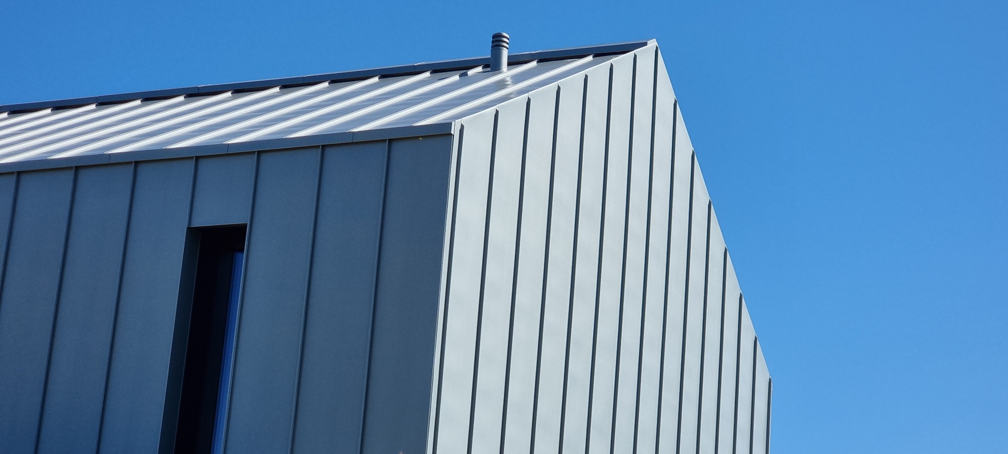 Nowoczesny dom z szarej blachy trapezowej: elewacja i dach w pionowe pasy, małe okno. Minimalistyczny design na tle błękitnego nieba.