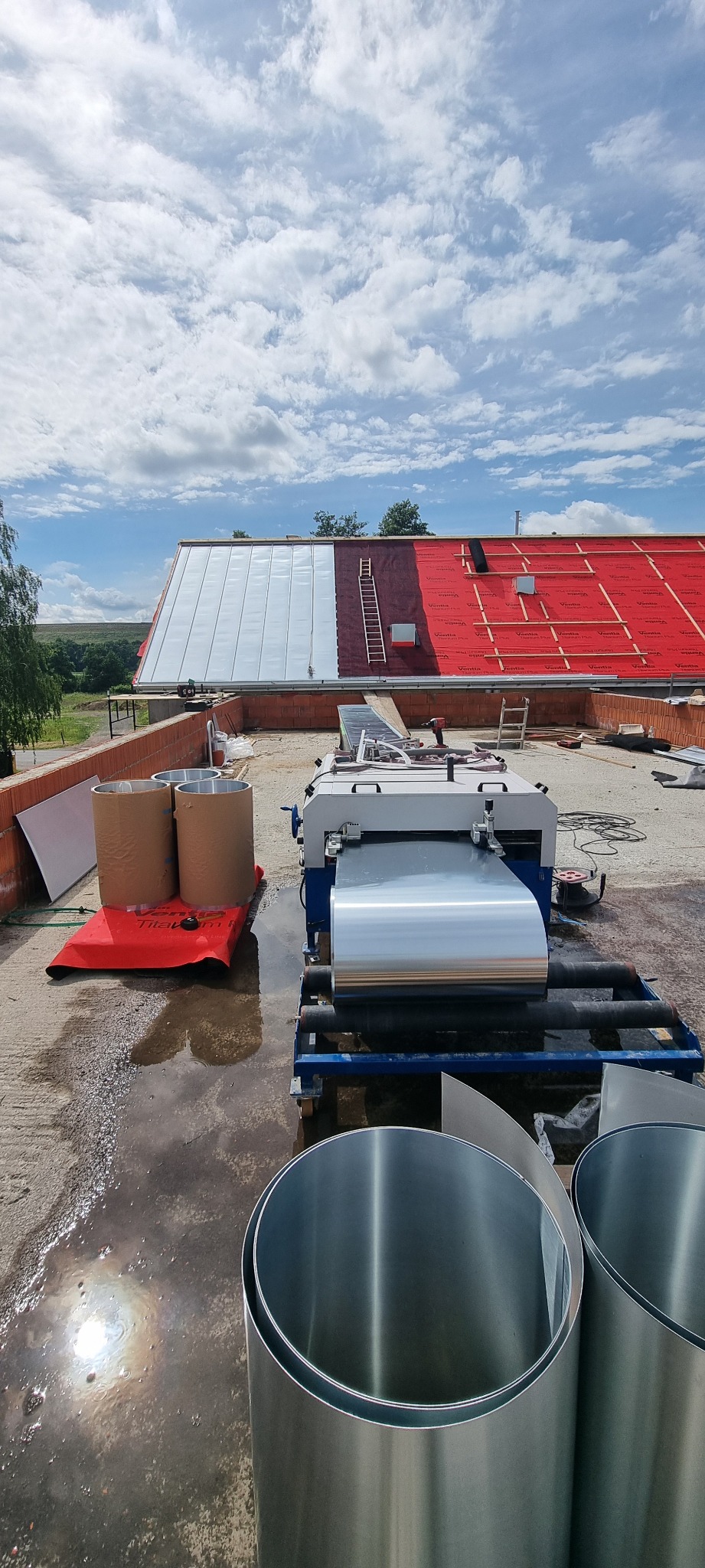 Dach w trakcie krycia blachą na rąbek stojący, widoczna maszyna do obróbki blacharskiej, drabina i częściowo położona membrana dachowa.