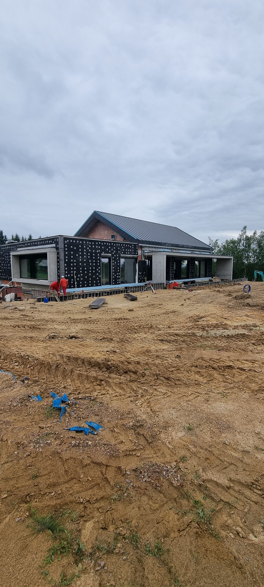 Nowoczesny dom w trakcie budowy: widoczne ocieplenie ścian zewnętrznych, pracownicy w czerwonych kombinezonach oraz fragment dachu z blachy na tle pochmurnego nieba.