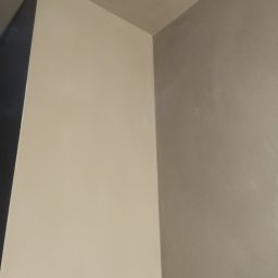 JAKUB JARÓG TYNKI MASZYNOWE - Gładka, beżowa ściana po prawej stronie styka się z szarą ścianą, tworząc narożnik. Widoczny fragment sufitu. Minimalistyczne wykończenie wnętrza.