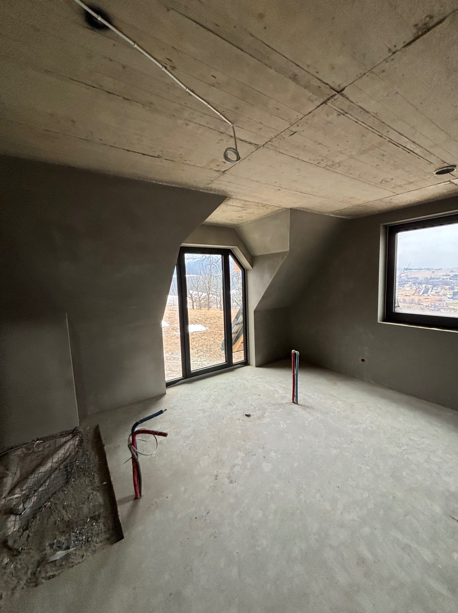 Pokój na poddaszu po tynkowaniu maszynowym ścian i sufitu. Widoczne okna z widokiem na krajobraz i wystające rury instalacyjne.