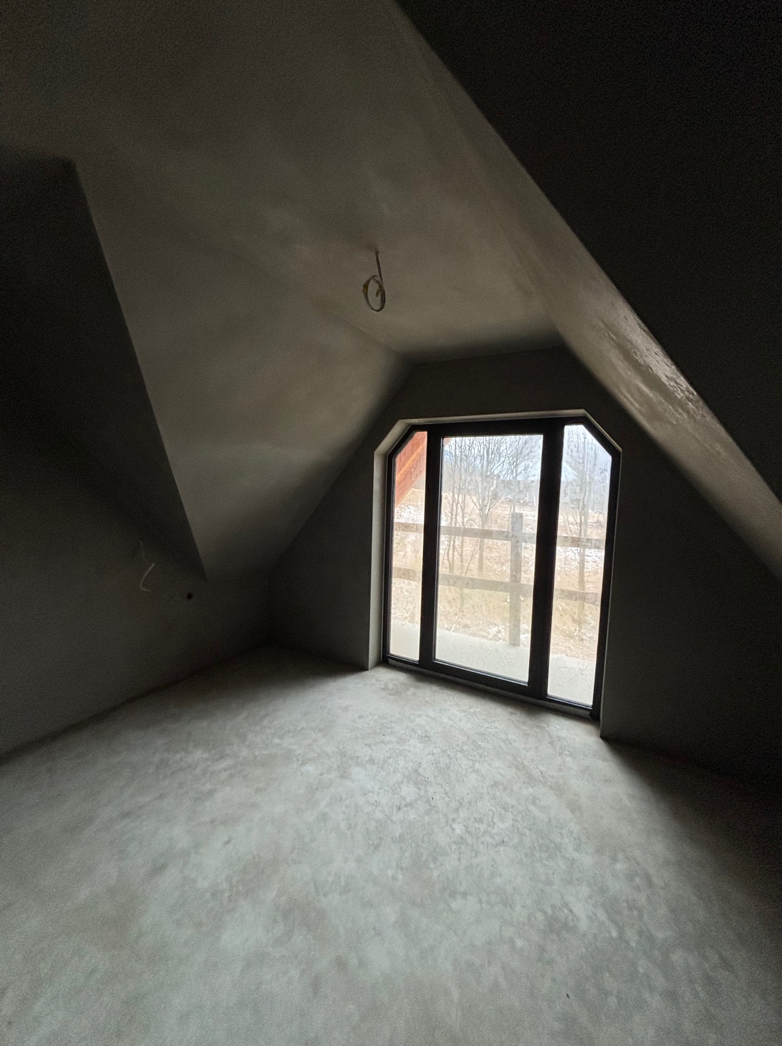 Pokój na poddaszu po tynkowaniu maszynowym. Widoczne gładkie, szare ściany i podłoga. Okno z widokiem na zewnątrz, przewody elektryczne wystające z sufitu.