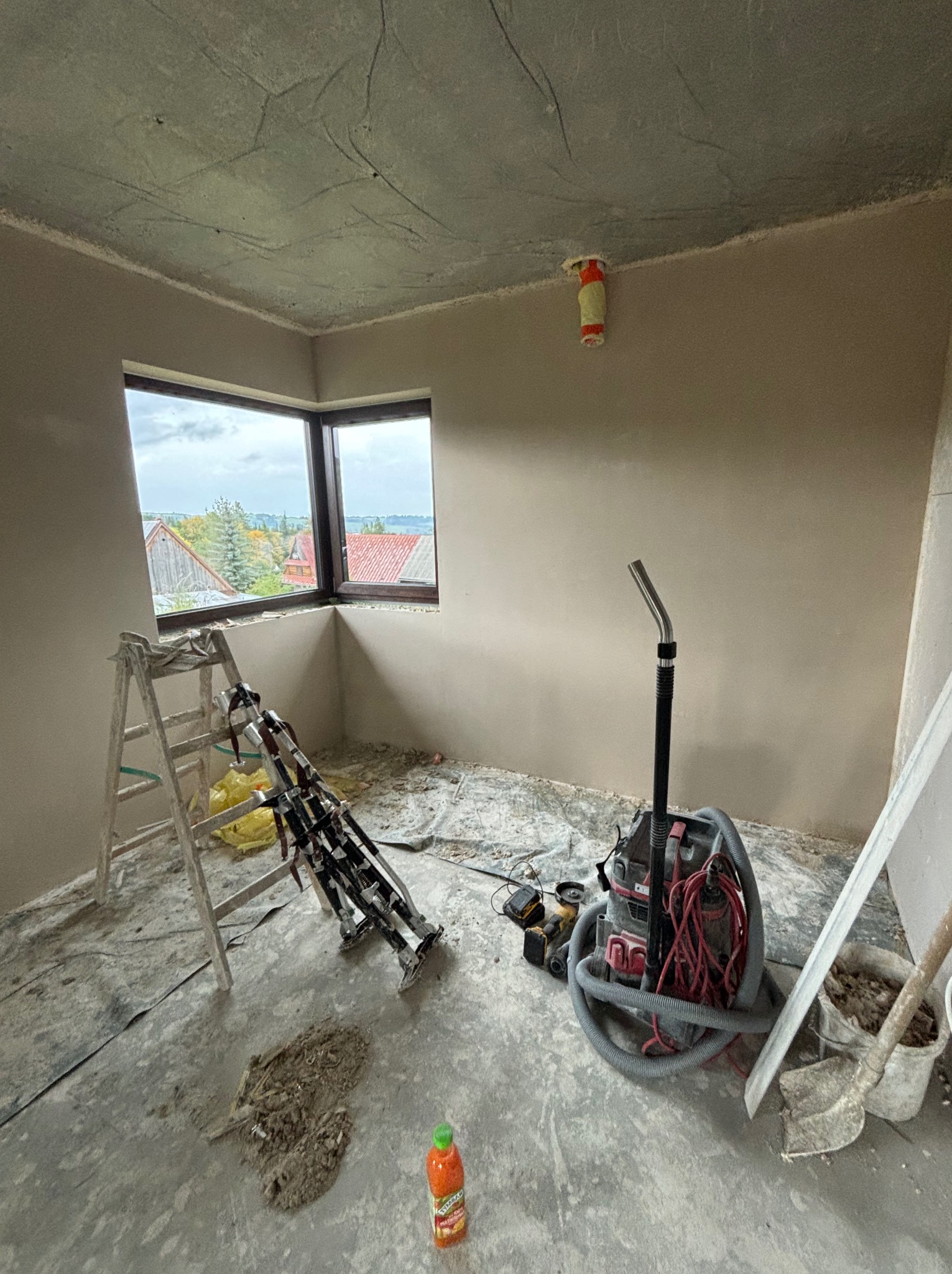 Pokój po pracach tynkarskich: świeże, gładkie ściany, drabina, narzędzia do tynkowania i odkurzacz przemysłowy. Widok przez okno na zabudowania i drzewa.